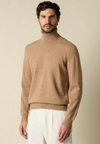 Khaki Rollkragenpullover aus einem weich gestrickten Stoff, mit einer figurbetonten Silhouette und gerippten Bündchen. Getragen mit hellen, gefalteten Hosen.