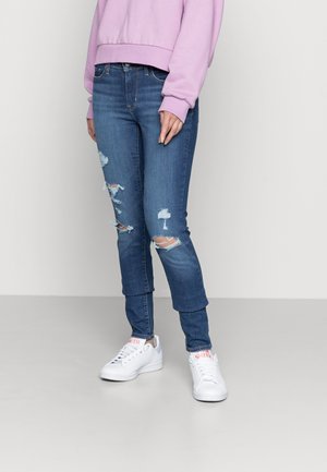 Jeans skinny en denim moyen, présentant des déchirures usées aux genoux. Associés à des baskets blanches et un sweat-shirt violet clair.
