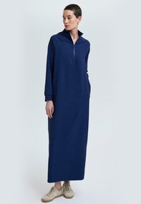 Femme debout dans une longue robe zippée navy à manches longues et poches latérales, portant des baskets beiges, sur un fond clair uni.