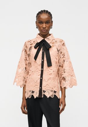 Žena v ružovej kvetinovej čipkovej blúzke s vlnitými okrajmi a čiernou viazačkou, v kombinácii s voľnými čiernymi nohavicami, stojaca na bielom pozadí.