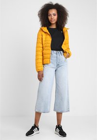 Gul pufferjakke med hette, svart t-skjorte, høyt liv lys blå jeans, og svarte sneakers med hvite detaljer, stående mot en ensfarget bakgrunn.