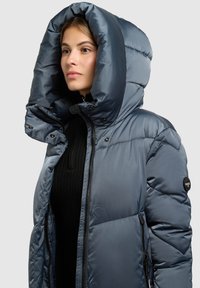 Tumesinist värvi toppatav jope, millel on suur kapuuts, ees tõmblukk ja soonikuga must sisekiht. Sile tekstuur õmblusega detailidega.