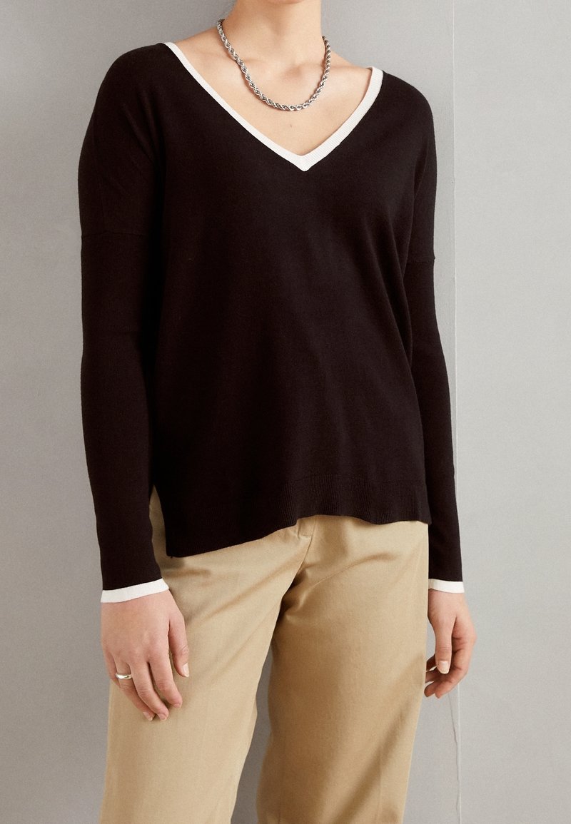 Personne portant un pull noir à col en V avec un bord blanc aux poignets et à l'encolure, un pantalon beige, un collier chaîne en argent et une bague à la main gauche.