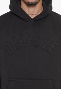 Sort hættetrøje lavet af blødt stof, med et hævet "DROPSIZE" logo tværs over brystet, samt kangaroo-lomme på forsiden.