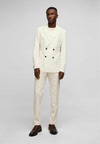 Hellbeige, doppelt geknöpfter Anzug mit dunklen Knöpfen, taillierte Passform und schmale Hosen. Kombiniert mit braunen Slippern. Glatte, gewebte Textur.