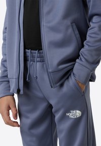 Plava sportska jakna na patent i odgovarajuće trenirke brenda The North Face, s glatkim materijalom, elastičnim pojasom i logotipom.