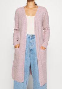 Donna che indossa un cardigan lungo di un rosa chiaro con apertura frontale e tasche laterali, una maglietta beige e jeans a vita alta di un blu chiaro.