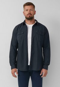 Camicia a maniche lunghe di colore blu navy scuro con due tasche sul petto, colletto e maniche ripiegate, indossata sopra una t-shirt bianca. Texture liscia.