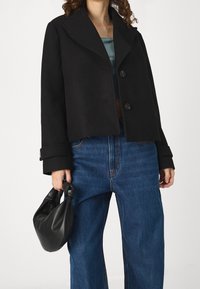 Černý zkrácený kabát z vlněné směsi s velkými klopami, dvojitým zapínáním na knoflíky a širokými rukávy. Kombinován s modrými džíny s vysokým pasem a černou taškou.