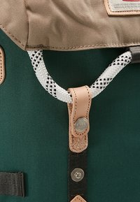 Sac à dos en tissu vert et beige avec cordon de serrage en corde blanche et noire, fixé par une sangle en cuir beige et des boutons-pression métalliques.