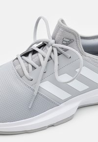 Harmaa urheilukenkä, jossa on verkkomateriaalia ylhäällä, litteät nauhat ja kolme valkoista raitaa. Pohja on valkoinen harmailla yksityiskohdilla ja siinä on tekstuuroitu muotoilu.