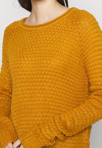 Närbild av en person som bär en strukturerad senapsgul stickad tröja med långa ärmar och rund hals. 