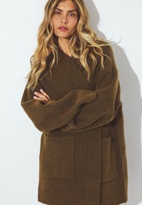 Cárdigan de punto oversize en verde olivo, con mangas largas, dos bolsillos frontales y detalles acanalados. Tejido texturizado con un aspecto suave.