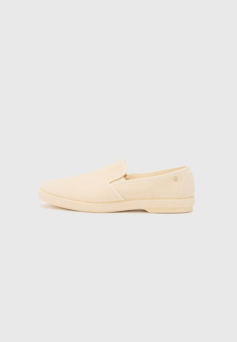 Chaussons en matériau beige clair, semblable à du feutre. Bout arrondi, texture lisse, design minimaliste et un détail de logo subtil près du talon.