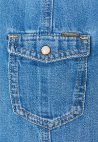 Cola de mezclilla azul claro con un botón a presión plateado, acentuada por costuras amarillas y una etiqueta negra que dice "Pepe Jeans."