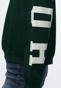 Manche de sweat-shirt vert foncé avec de grandes lettres blanches en bloc et un jean en denim bleu visible à la taille et à la hanche.