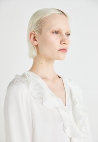 Blusa bianca con scollo a V profondo, caratterizzata da un bordo arricciato lungo il colletto. Texture liscia e setosa, con maniche lunghe visibili.