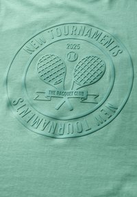 Tričko v mátově zelené barvě s vystouplým potiskem tenisových raket, nápisy "NOVÉ TURNAJI" a "TENISOVÝ KLUB 2025" v kruhovém designu.
