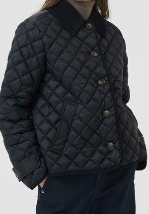 Woman wearing a black quilted jacket with button closure, angled pockets, and a dark corduroy collar over a brown turtleneck.