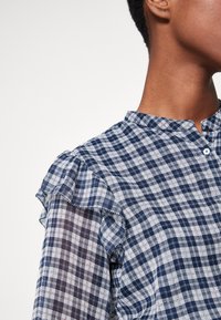 Blusa a quadri blu e bianchi con dettagli arricciati sulle spalle, dotata di una chiusura con bottoni e realizzata in un tessuto leggero.