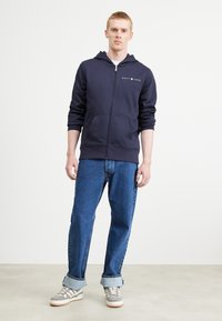 Felpa con zip color navy e tasche a marsupio, logo bianco sul petto, indossata con jeans blu a maniche corte e sneaker grigie. Tessuto di cotone liscio.