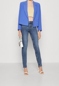 Blazer bleu raccourci porté sur un top court beige, associé à un jean skinny bleu foncé et des sandales blanches à talons blocs, tenant un petit sac blanc.