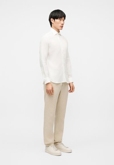 Άνδρας που στέκεται φορώντας λευκό πουκάμισο μακρυμάνικο με κουμπιά, μπεζ παντελόνι και λευκά αθλητικά παπούτσια σε απλό λευκό φόντο.