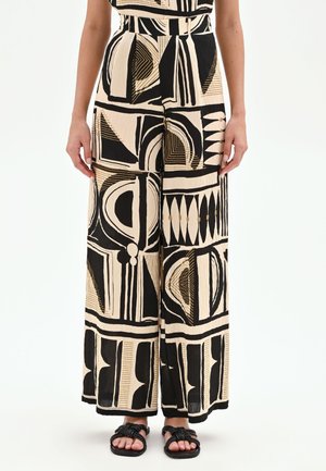 Femme portant un pantalon taille haute à jambes larges avec un imprimé géométrique abstrait noir et beige et des sandales plates noires.