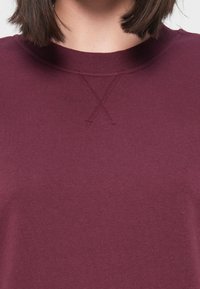 Vue partielle d'une personne portant un pull col rond violet foncé avec un détail "X" cousu sous le col.