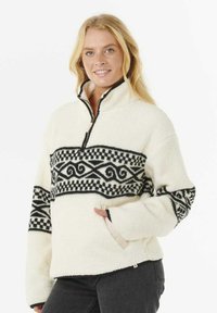Fleece pullover, crèmekleurig met een zwart geometrisch patroon. Heeft een ritskraag, zijzakken en een zachte textuur.