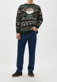 Pull en maille verte avec un motif festif comprenant un personnage de dessin animé, le texte "QUE LA FORCE DE JOIE SOIT AVEC TOI", et des motifs contrastés rouges et blancs.