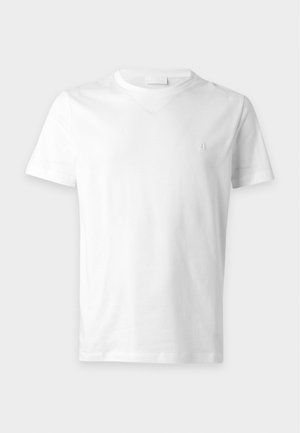 Jeune homme debout portant un t-shirt blanc superposé à une chemise verte, un jean bleu déchiré et des mocassins noirs sur un fond uni.