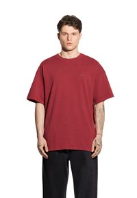 Bordowa bawełniana koszulka z okrągłym dekoltem, w oversize'owym kroju, z krótkim rękawem i delikatnym detalem z logo na klatce piersiowej, noszona z czarnymi spodniami.