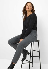 Zwarte langarmshirt gecombineerd met grijs gepatternde broek. Schoenen zijn zwarte enkelboots. Model zit op een zwarte kruk tegen een witte achtergrond.