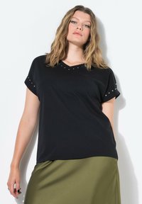 Žena s vlnitými blond vlasy v černém tričku s cvočky s krátkým rukávem a olivově zelené sukni na bílém pozadí.