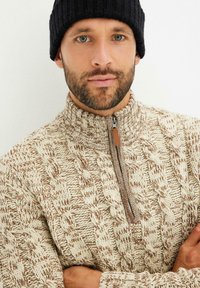 Maglione lavorato a maglia color crema con motivi a treccia marroni, caratterizzato da un collo alto e una mezza cerniera. Un berretto a coste nero è indossato come accessorio.