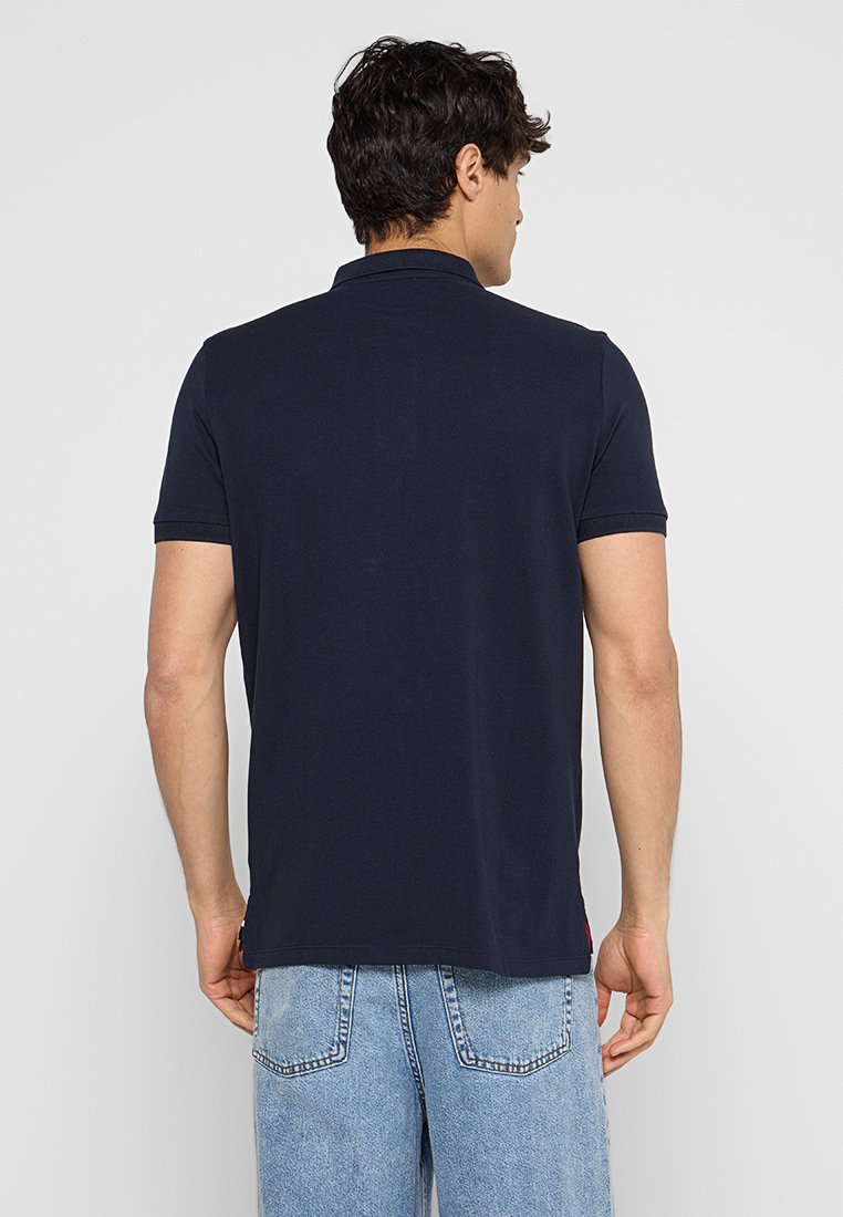 Polo shirt bleu marine avec col classique et manches courtes. Fabriqué en tissu lisse, il présente un ourlet droit et un détail de logo subtil sur le côté.