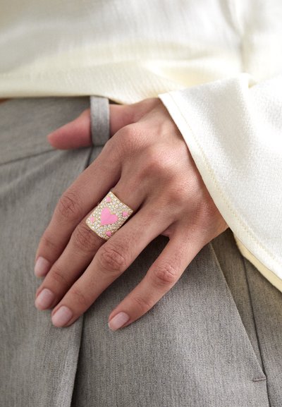 Anillo dorado con acentos en forma de corazón rosa y rhinestones claros, de forma rectangular, usado en una mano descansando sobre una tela gris. Textura suave.
