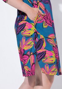 Wielobarwna sukienka z niebieskiego materiału z dynamicznymi różowymi, żółtymi i fioletowymi wzorami kwiatowymi. Zawiera boczną kieszeń oraz rozcięcie dla wygody.