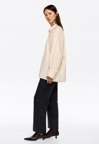 Mujer de pie de lado, vistiendo una camisa suelta beige, pantalones negros de pierna ancha y botines negros de tacón, contra un fondo blanco.