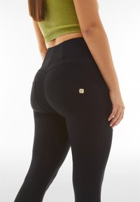 Leggings neri a vita alta realizzati in materiale liscio, dotati di tasche posteriori curve e un piccolo accento con logo dorato.