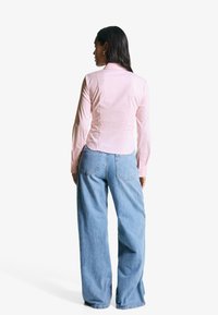 Camicia a maniche lunghe rosa chiaro con un design attillato e orlo curvo, abbinata a jeans in denim azzurro chiaro, larghi, con taglio classico.