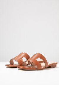 Brown leather sandals with a flat heel, featuring cut-out detailing and contrasting stitching, presenting a smooth texture and minimalist design.