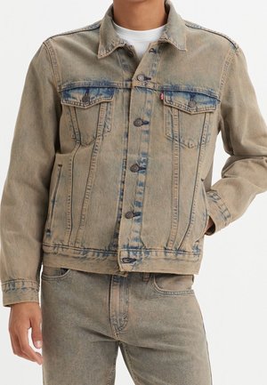 Mann trägt eine beige Jeansjacke und passende Jeans mit Knopfverschluss und Brusttaschen, steht vor einem einfarbigen Hintergrund.