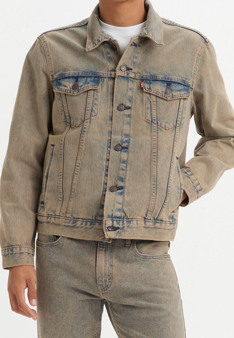Homme portant une veste en jean beige et un jean assorti avec des fermetures à boutons et des poches poitrine, debout devant un fond uni.