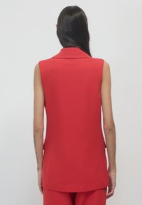 Blazer rosso senza maniche con ampio colletto, texture liscia, vestibilità sartoriale e lati con spacco. Tessuto leggero, design pulito, minimalismo nei dettagli.