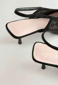 Čierne sandále na vysokom podpätku s čipkovým vzorom a ružovou stielkou. Majú štvorecový špičku, štíhly členkový remienok a krátke blokové podpätky.