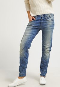 Blå denimjeans med tapered passform. Har bleknings- och whiskeringdetaljer. Bärs med en krämfärgad stickad tröja och vita sneakers.
