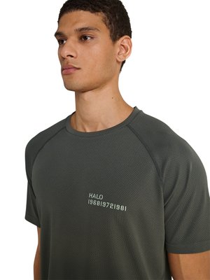 Junger Mann trägt ein dunkelgraues kurzärmeliges strukturiertes Hemd mit der Aufschrift „HALO 196819721981“ auf der Brust, leicht nach links gewandt.