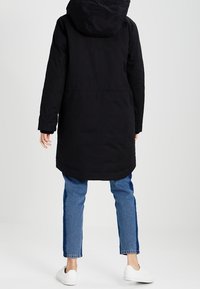 Zwarte lange jas met een capuchon, met een rechte snit en zakken. Gepaard met blauwe jeans en witte sneakers. Eenvoudig en functioneel ontwerp.